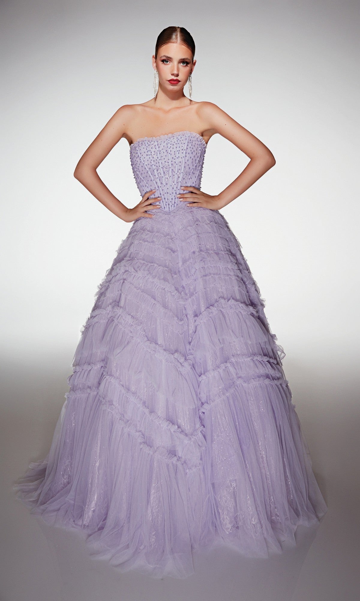 A woman stands with hands on hips, wearing the Alyce 62208 long prom dress—a strapless ballgown with a tulle ruffle skirt and beaded embellishments. This floor-length lavender gown exudes elegance and grace.